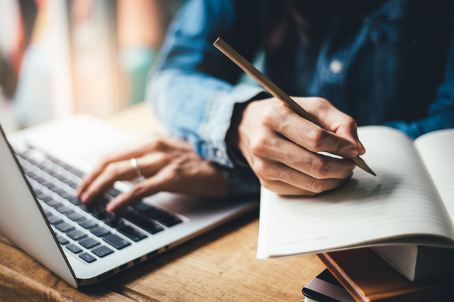 man writing in notepad next to laptop